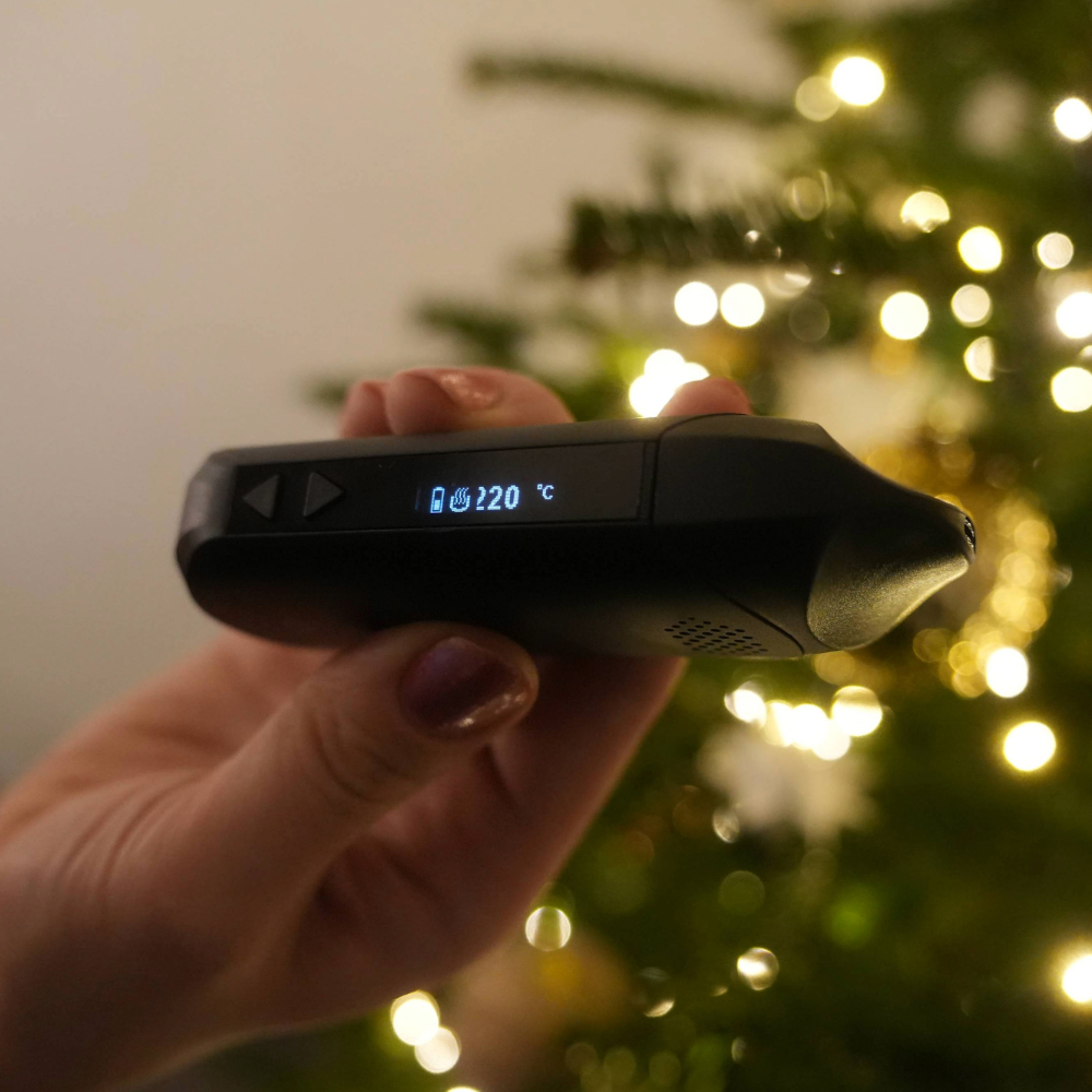 Nahaufnahme des Flowermate The Cap Pro Displays mit Anzeige der Temperatur auf 220°C, gehalten vor einer weihnachtlichen Kulisse.