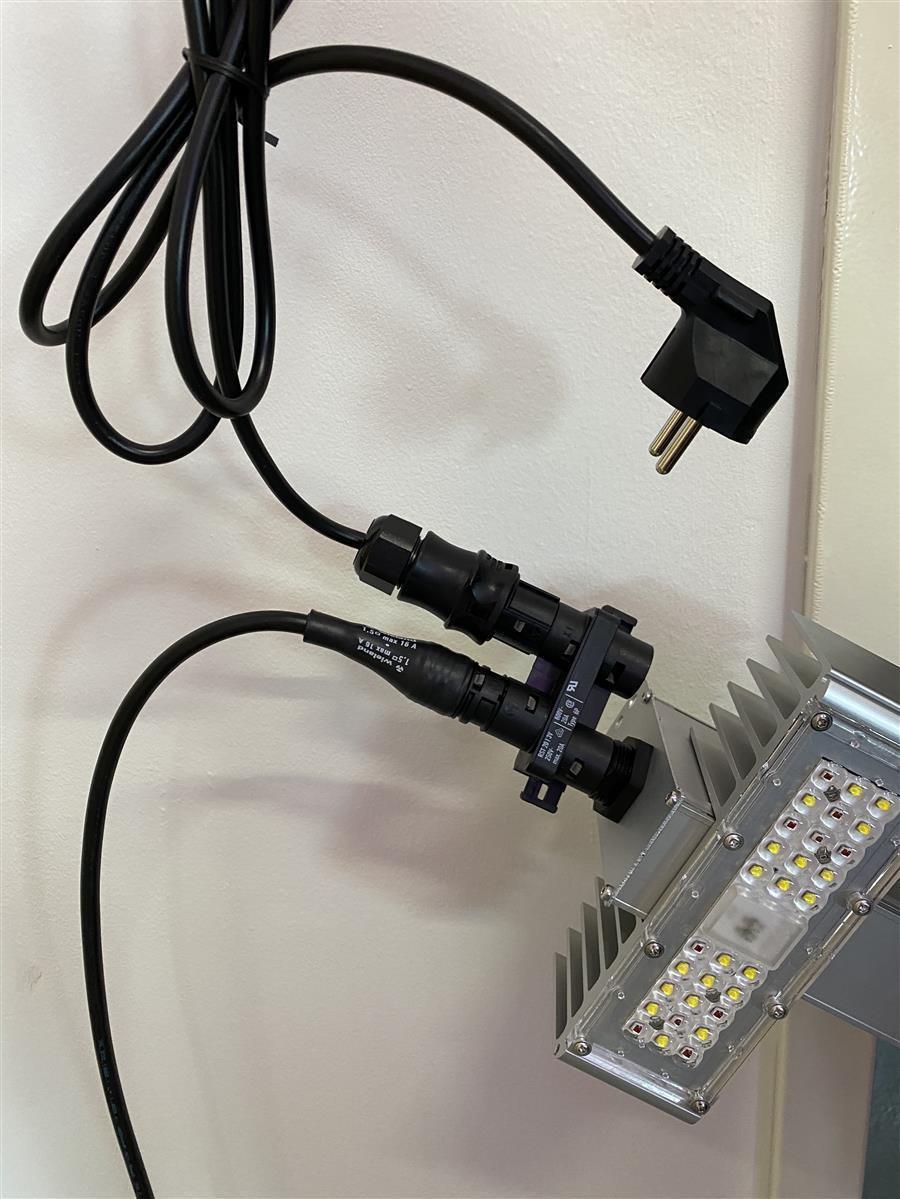 Schwarzes Netzkabel mit Schukostecker und verbundenem Adapter, an eine LED-Lichtleiste angeschlossen, gegen eine Wand montiert.