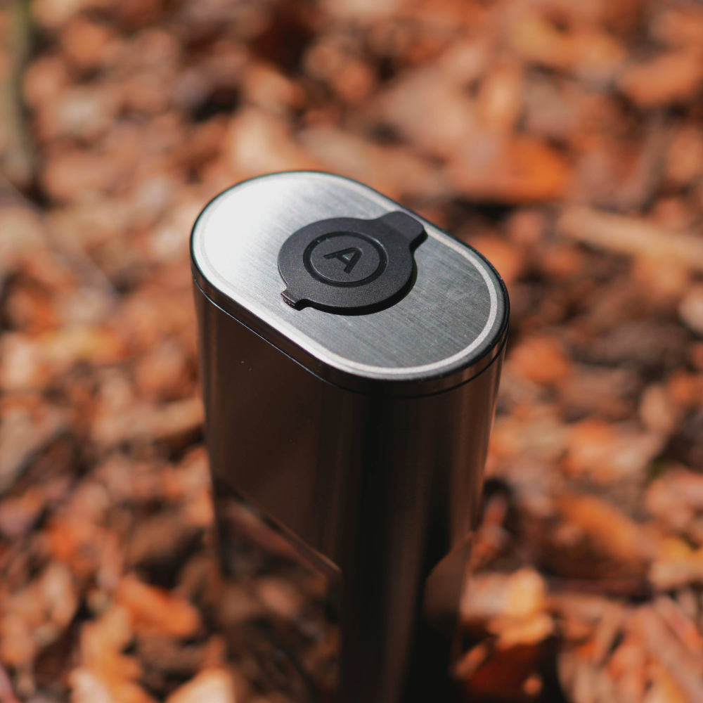 Nahaufnahme der gebürsteten Metalloberfläche eines tragbaren Vaporizers mit Deckel und 'A'-Logo im herbstlichen Wald